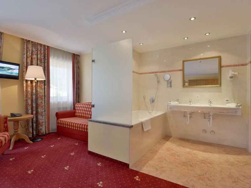 Hotelzimmer mit rotem Teppich, Sitzecke und offenem Badezimmer mit zwei Waschbecken.