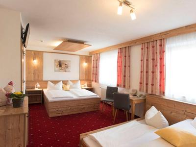 Camera d'hotel luminosa con letto matrimoniale, letto singolo, moquette rossa e finestre con tende arancioni.