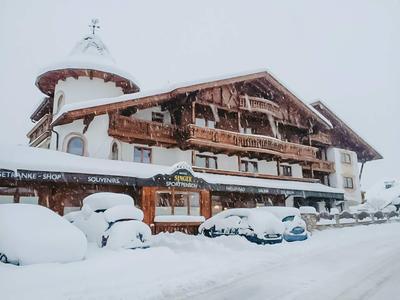 Ein verschneites Hotel mit Holzbalkonen und mehreren schneebedeckten Autos vor dem Gebäude.