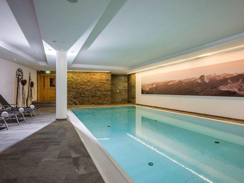 Innenraum eines Schwimmbads mit blauem Wasser, weißen Wänden, Liegestühlen und großem Landschaftsgemälde.