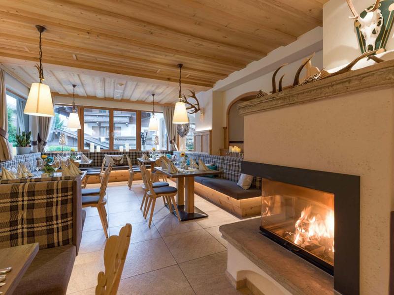 Gemütlicher Gastraum mit Holzmöbeln, Kaminfeuer, großen Fenstern und warmem Licht.