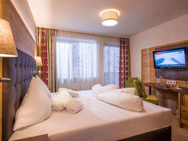 Gemütliches Hotelzimmer mit Doppelbett, Holzmöbeln, Fernseher und großen Fenstern mit Vorhängen.