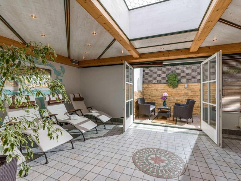 Heller Ruheraum mit Liegestühlen, Glasdach und offener Tür zu sonniger Terrasse mit Sitzgruppe.