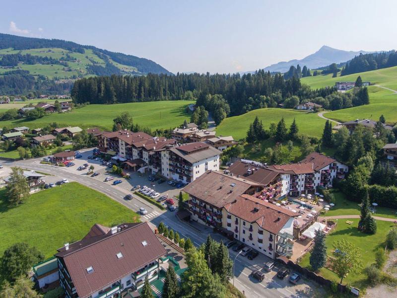 Dorf mit mehreren Gebäuden und Parkplätzen, umgeben von grünen Wiesen und bewaldeten Hügeln.