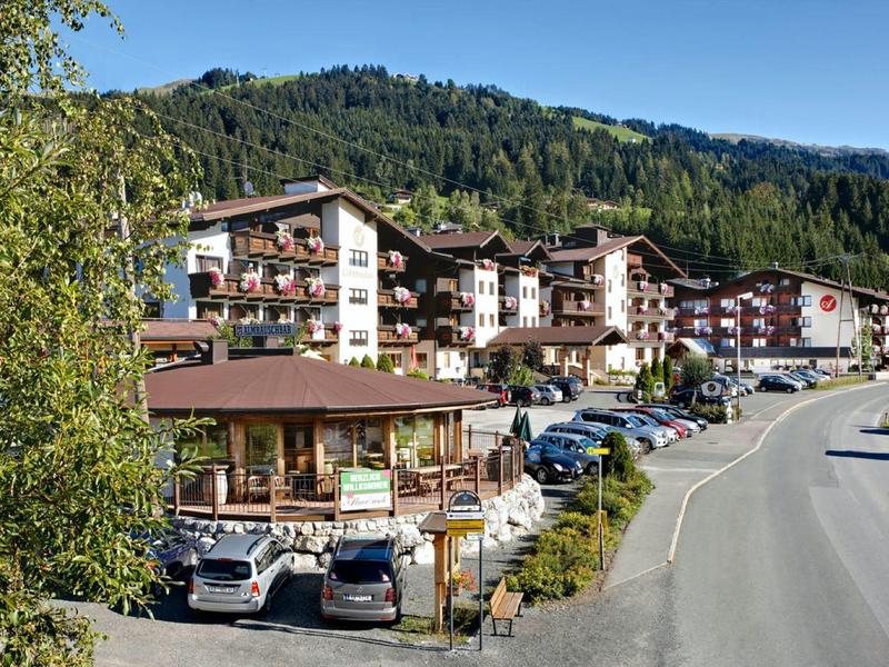 Dorf mit traditionellen Gebäuden, Autos auf Parkplätzen, Berglandschaft und blauem Himmel im Hintergrund.