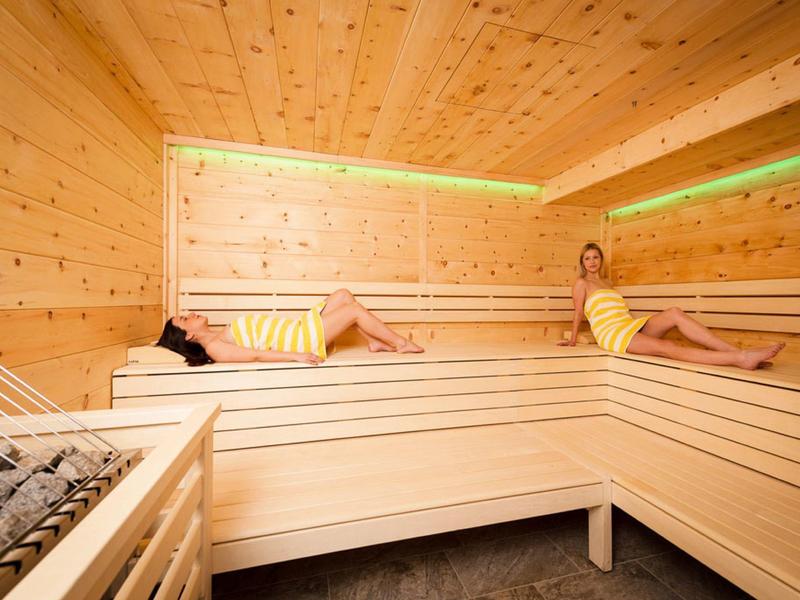Sauna aus hellem Holz mit zwei liegenden Personen auf mehrstufigen Bänken und grüner LED-Beleuchtung.