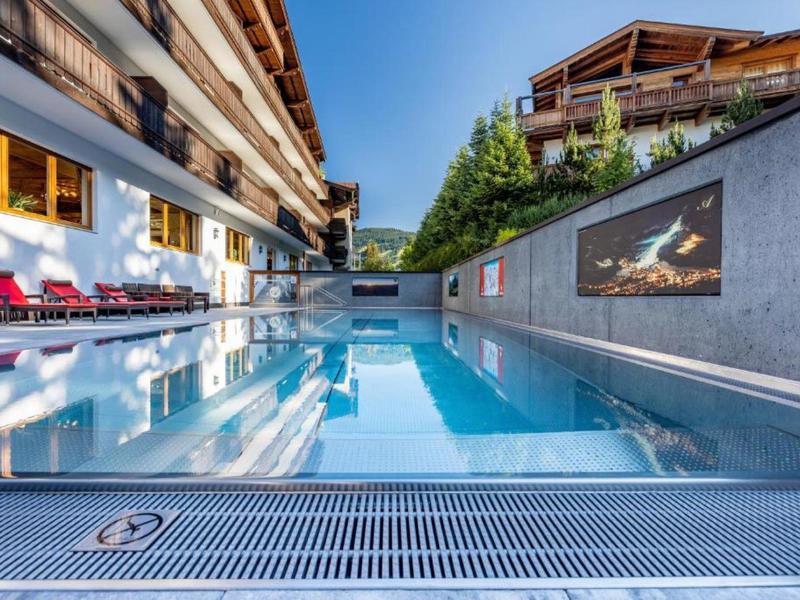 Moderner Außenpool mit Liegestühlen und Holzchalets bei klarem Himmel, umgeben von grünen Pflanzen.