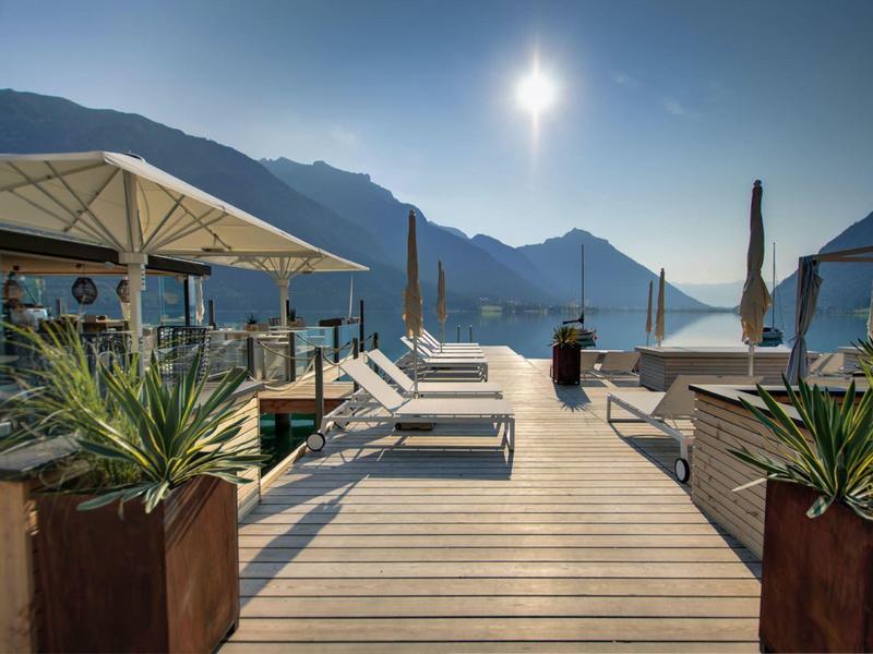 Holzterrasse mit Liegestühlen, Sonnenschirmen und Pflanzen, umgeben von Bergen und klarem Himmel.