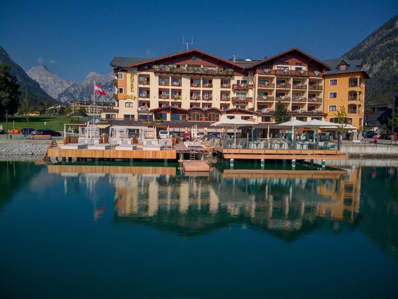 Großes Hotel an Bergsee, reflektiert im ruhigen Wasser, umgeben von grünen Hügeln und Bergen.