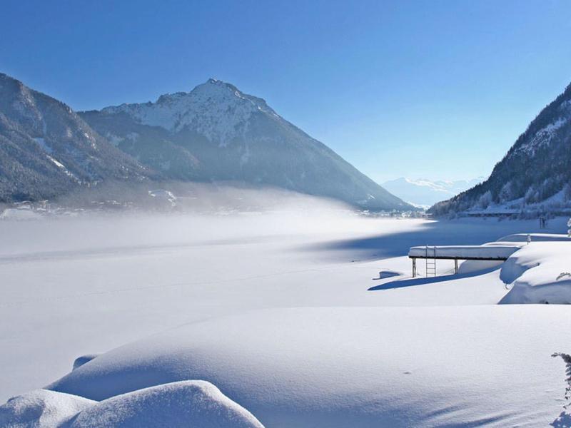 Verschneites Tal mit Bergkulisse, blauem Himmel und Nebel auf dem gefrorenen See.