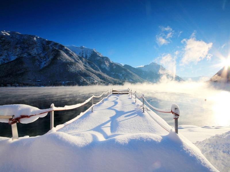 Winterlicher Steg im See, umgeben von schneebedeckten Bergen und strahlend blauem Himmel.