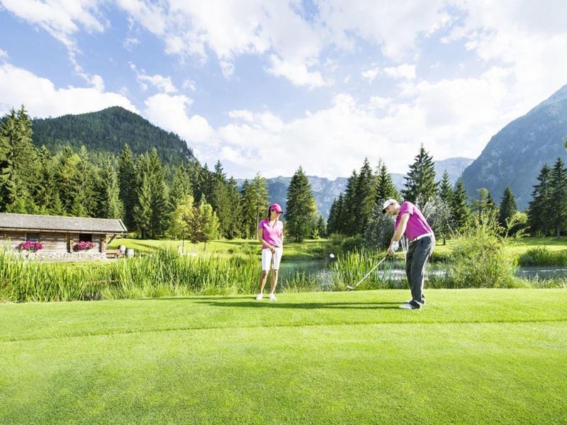 Zwei Golfer auf grünem Golfplatz vor Bergkulisse mit Bäumen und blauem Himmel.