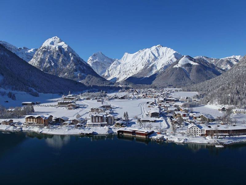 Dorf mit verschneiten Häusern an See, umgeben von schneebedeckten Bergen unter klarem Himmel.