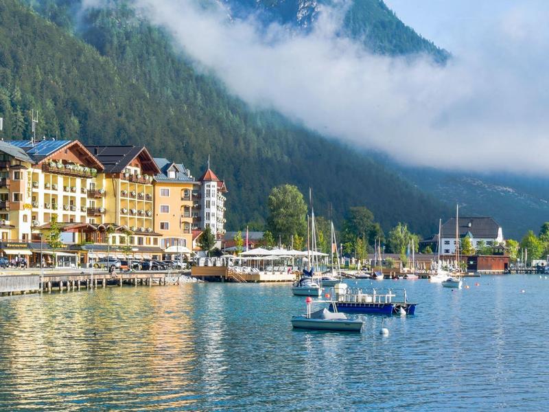 Bergsee mit Segelbooten und bunten Häusern am Ufer, umgeben von bewaldeten Bergen und Wolken.