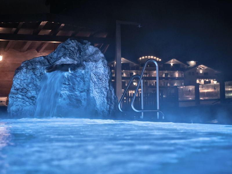 Beleuchtetes Hallenbad mit Wasserfall und Treppe, Umgebung mit warmem Licht in der Nacht.