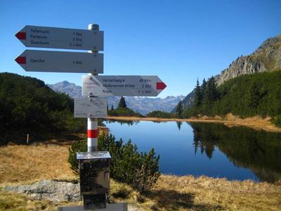 Hiking signpost by a calm lake with mountains in the background under a clear sky.