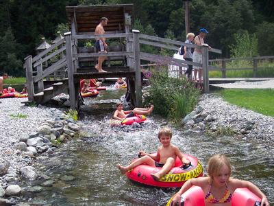 Children play with inflatable boats in a clear stream beside a wooden bridge in a green landscape.