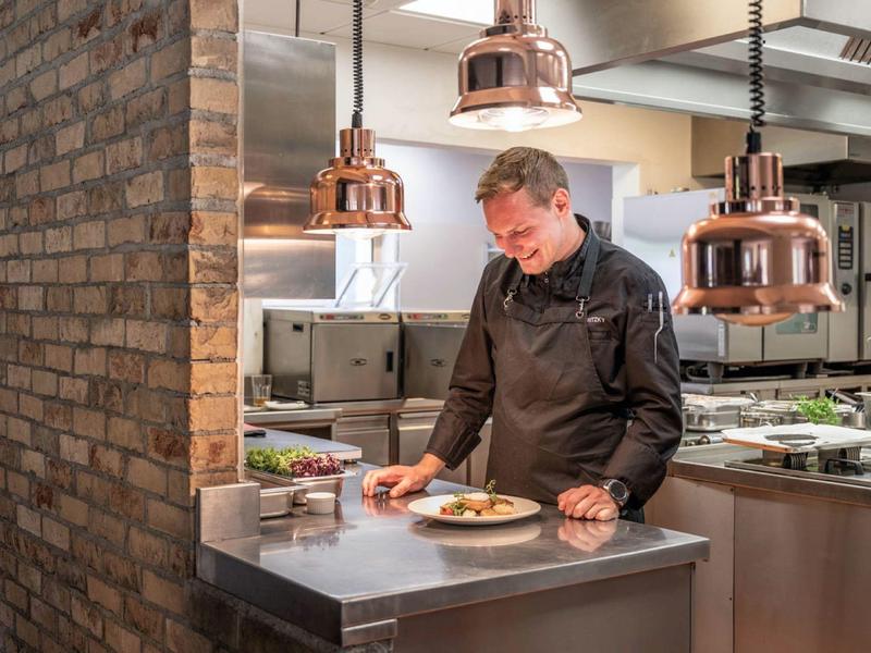 Chef inspects a plated dish in a modern hotel kitchen with copper lighting.