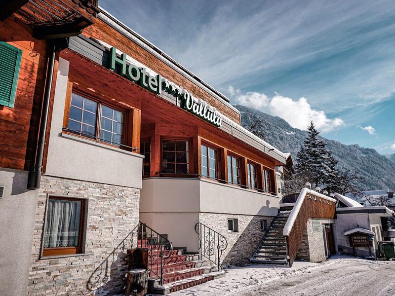 Holzhaus-Hotel im Bergstil mit Schnee, Treppe, grünen Fensterläden und blauem Himmel im Hintergrund.
