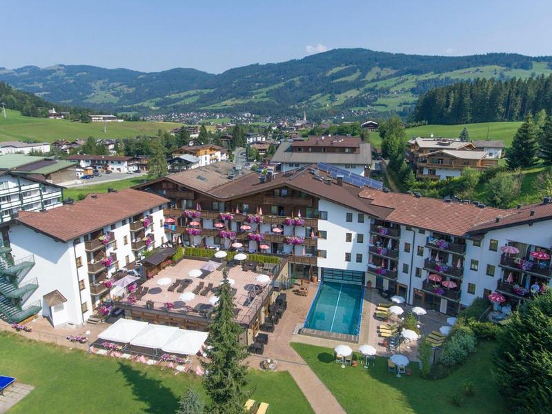 Großes Hotel mit Innenhof, Außenpool, Wiesen, Bergen im Hintergrund und blauem Himmel.