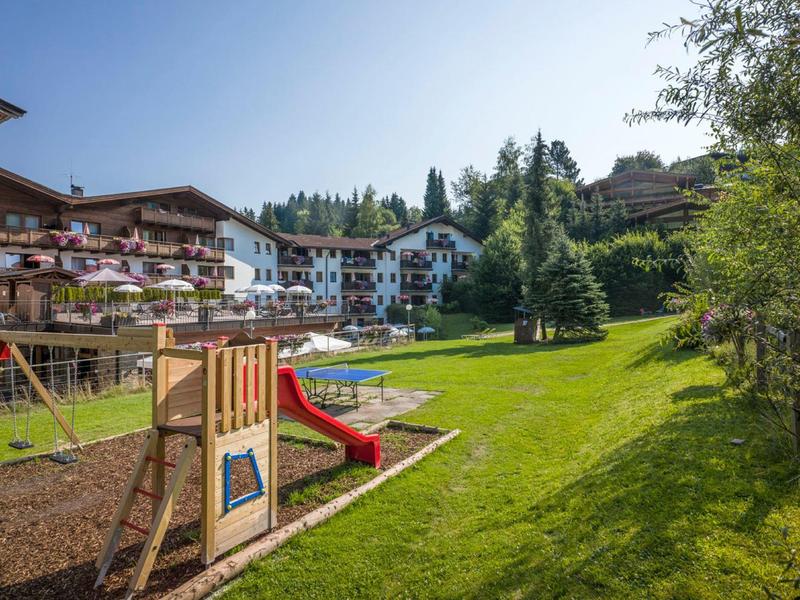 Spielplatz mit Rutsche und Schaukeln vor mehrstöckigen Häusern und grüner Wiese bei klarem Himmel.