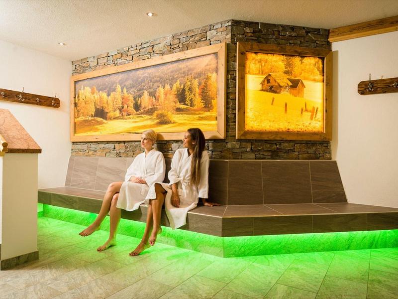 Zwei Frauen in weißen Bademänteln sitzen auf einer Bank mit grünem LED-Licht und herbstlichen Bildern.