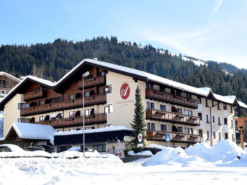 Hotel im verschneiten Bergdorf mit Holzbalkonen und Tannen vor bewaldeten Hügeln
