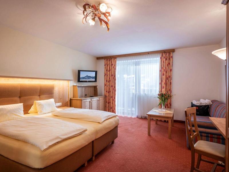 Hell eingerichtetes Hotelzimmer mit Doppelbett, Teppichboden, Schreibtisch und Balkon mit Vorhängen.