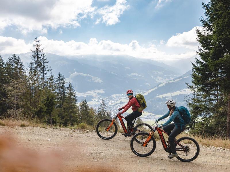 Zwei Radfahrer auf einem Bergpfad mit Bäumen und Wolken am Himmel.