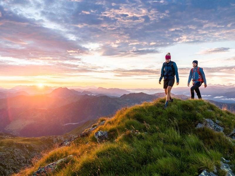 Zwei Wanderer stehen auf grün bewachsenem Hügel, während die Sonne hinter Bergen untergeht.