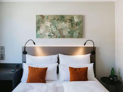 Moderní dvojlůžko s bílým povlečením a dvěma oranžovými polštáři v hotelovém pokoji.