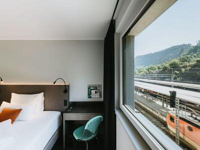 Moderní hotelový pokoj s jednolůžkem, psacím stolem a velkým oknem s výhledem na vlak a hory.