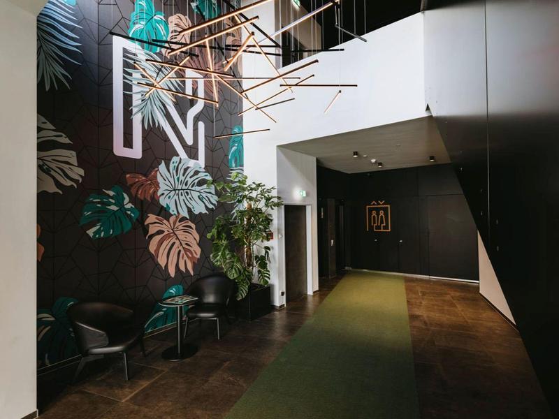 Moderní hotelová chodba s tmavou zdí a velkými rostlinnými vzory, osvětlená neonovým světlem.