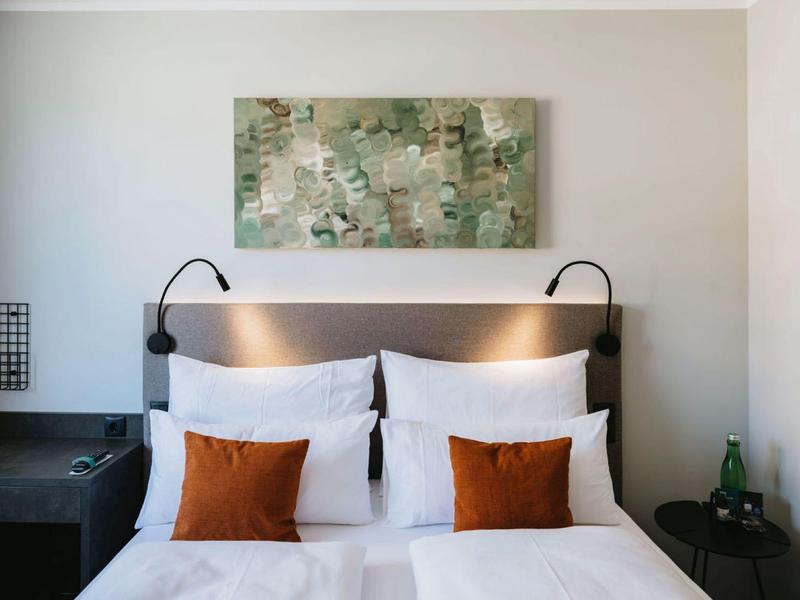 Moderní dvojlůžko s bílým povlečením a dvěma oranžovými polštáři v hotelovém pokoji.