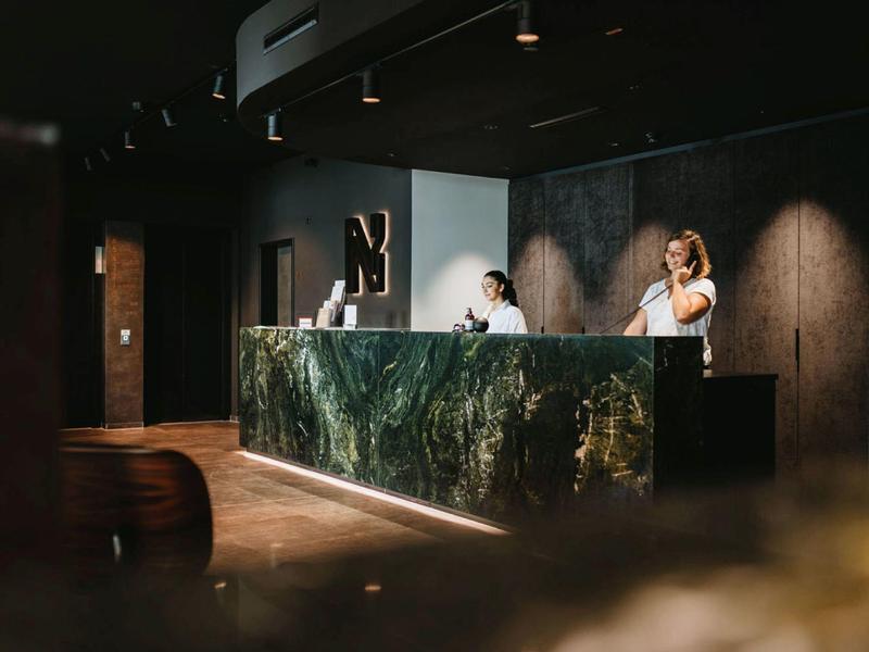 Recepce v moderním hotelu s tmavým, stylovým designem a dvěma zaměstnanci u přepážky.