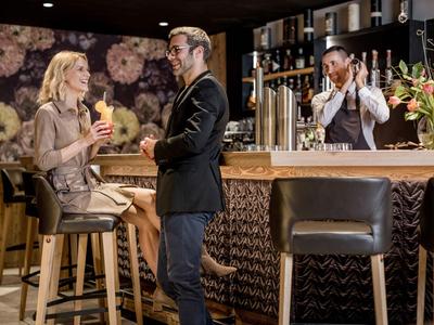 Hotelbar mit Frau und Mann, Barkeeper im Hintergrund und Blumendekoration am Tresen.