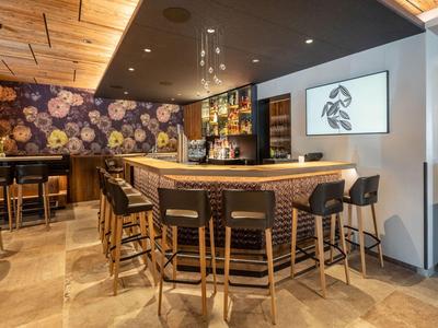 Moderner Hotelbarbereich mit Holzdecke, Blumenmuster an der Wand und Stühlen aus geflochtenem Material.