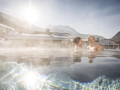 Zwei Personen entspannen in einem beheizten Außenpool mit Bergblick bei Sonnenschein.
