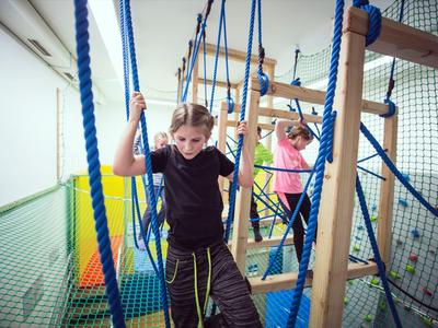 Kinder navigieren durch ein Klettergerüst mit Seilen in einem Indoor-Spielbereich.