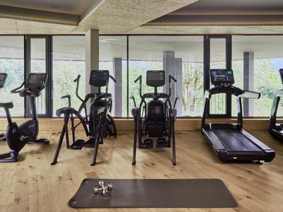 Fitnessraum mit Cardio-Geräten und Yogamatte vor großen Fenstern mit Blick auf Grünflächen.