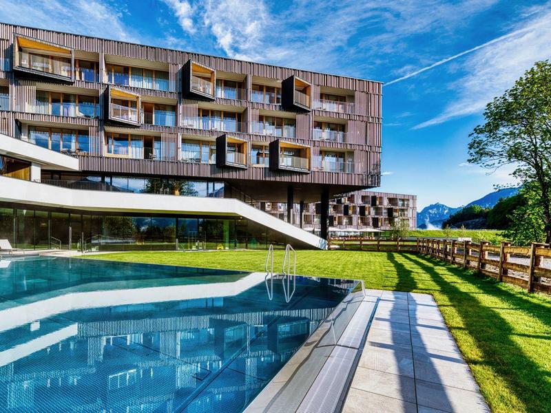 Edificio di hotel moderno con piscina all'aperto e vista sulle montagne sotto un cielo soleggiato.