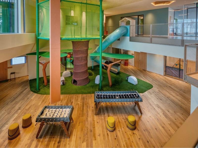 Area giochi interna moderna con struttura per arrampicata, scivolo e calcio balilla in un salone luminoso.