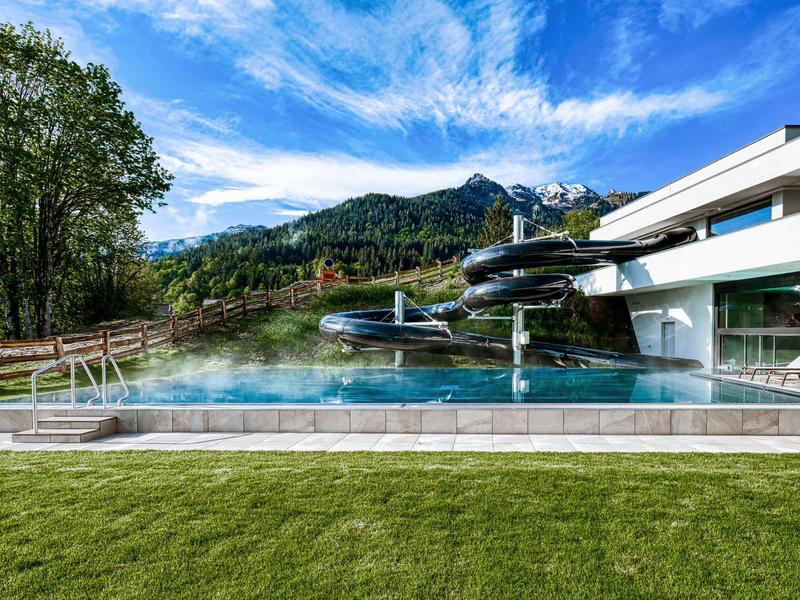 Piscina esterna moderna con scivolo davanti a un paesaggio montano e cielo azzurro