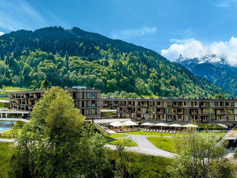 Hotel moderno in paesaggio montano verde con prati fioriti e cielo nuvoloso.