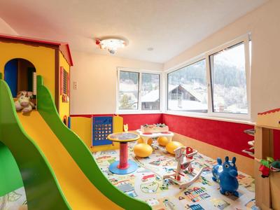 Heller Kinderraum mit großem Fenster, buntem Teppich, grün-gelber Rutsche und Spielzeug.