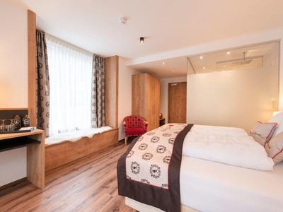 Helles Hotelzimmer mit großem Bett, Holzfußboden, Fensterbank und rotem Sessel am Fenster.