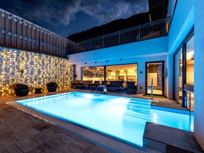 Moderner Außenpool mit blauer Beleuchtung, Sitzbereich, beleuchteter Wand und Bergkulisse bei Nacht.