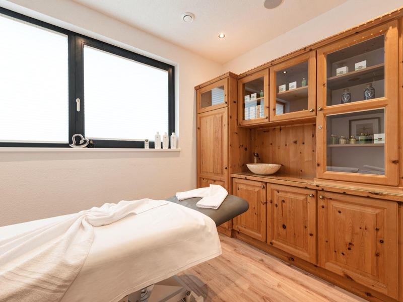 Heller Raum mit Massageliege, Holzmöbeln und großem Fenster. Gemütliche, ruhige Atmosphäre.