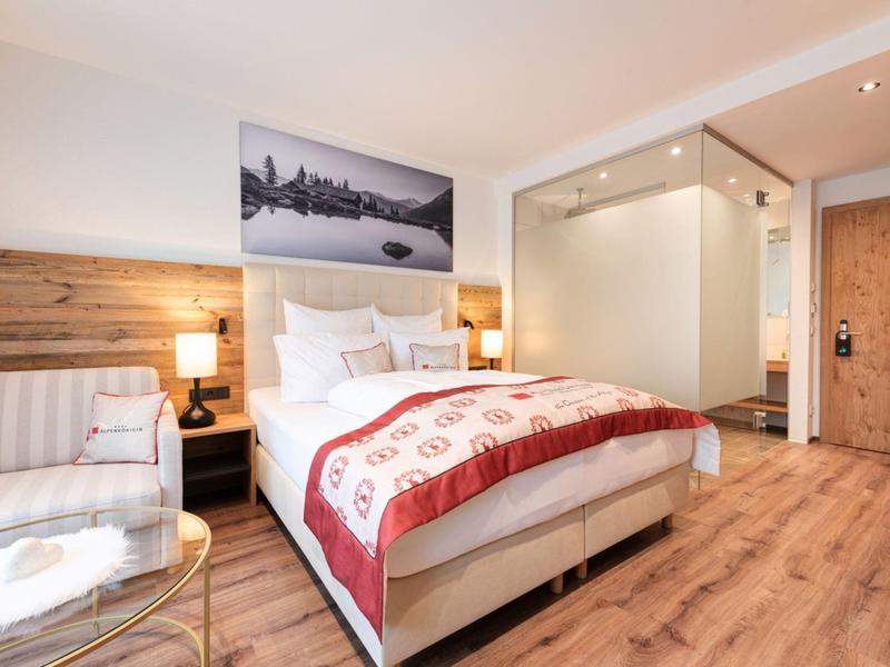 Modernes Schlafzimmer mit großem Bett, Holzfußboden, Glas-Duschkabine und gemütlicher Beleuchtung.