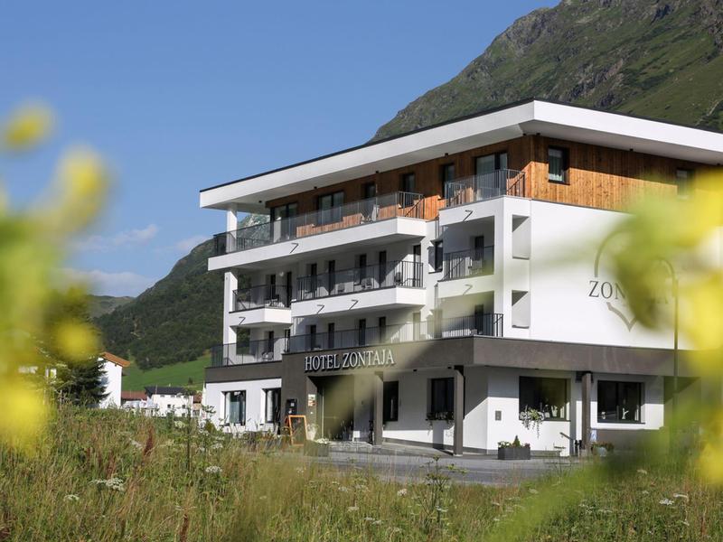 Modernes, mehrstöckiges Hotelgebäude mit Balkonen, umgeben von grüner Wiese und Bergen im Hintergrund.